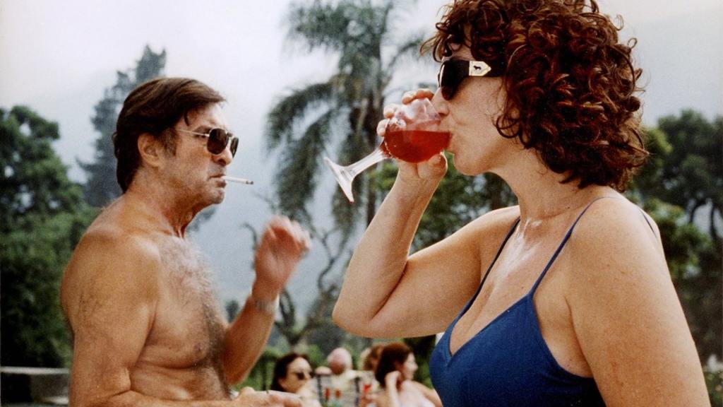 A man and a woman wearing sunglasses smoke and drink by the pool in a scene from Lucrecia Martel's La Ciénaga (2001)