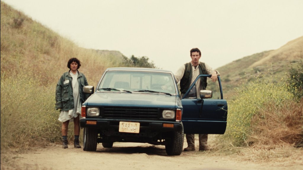 Alia Shawkut and Callum Turner stand on either side of a car parked on a deserted road in Atropia, reviewed on Single Take with Daniel Barnes.