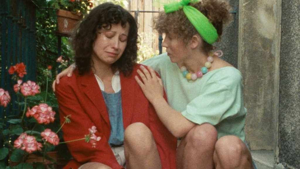 A crying woman in a red jacket is comforted by a woman dressed all in green in a scene from The Green Ray (1986).