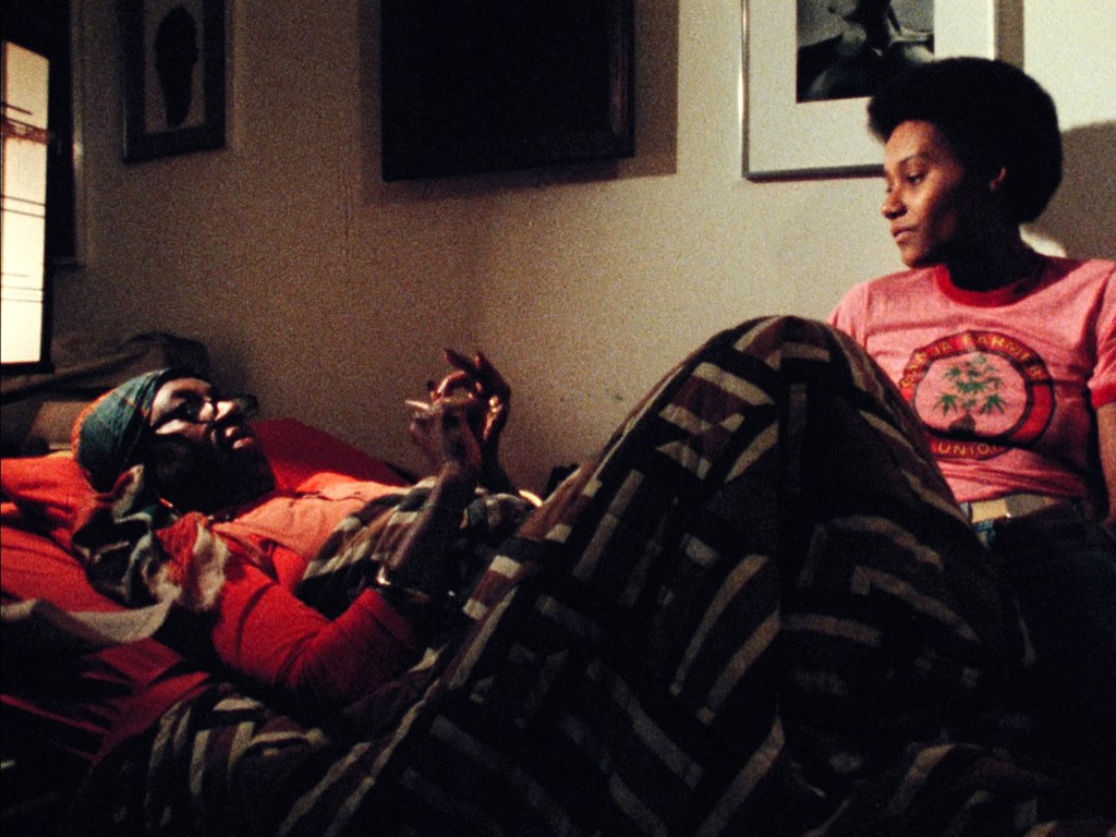 Two black women, one lying down in bed, one sitting up, have a conversation in a scene from Born in Flames